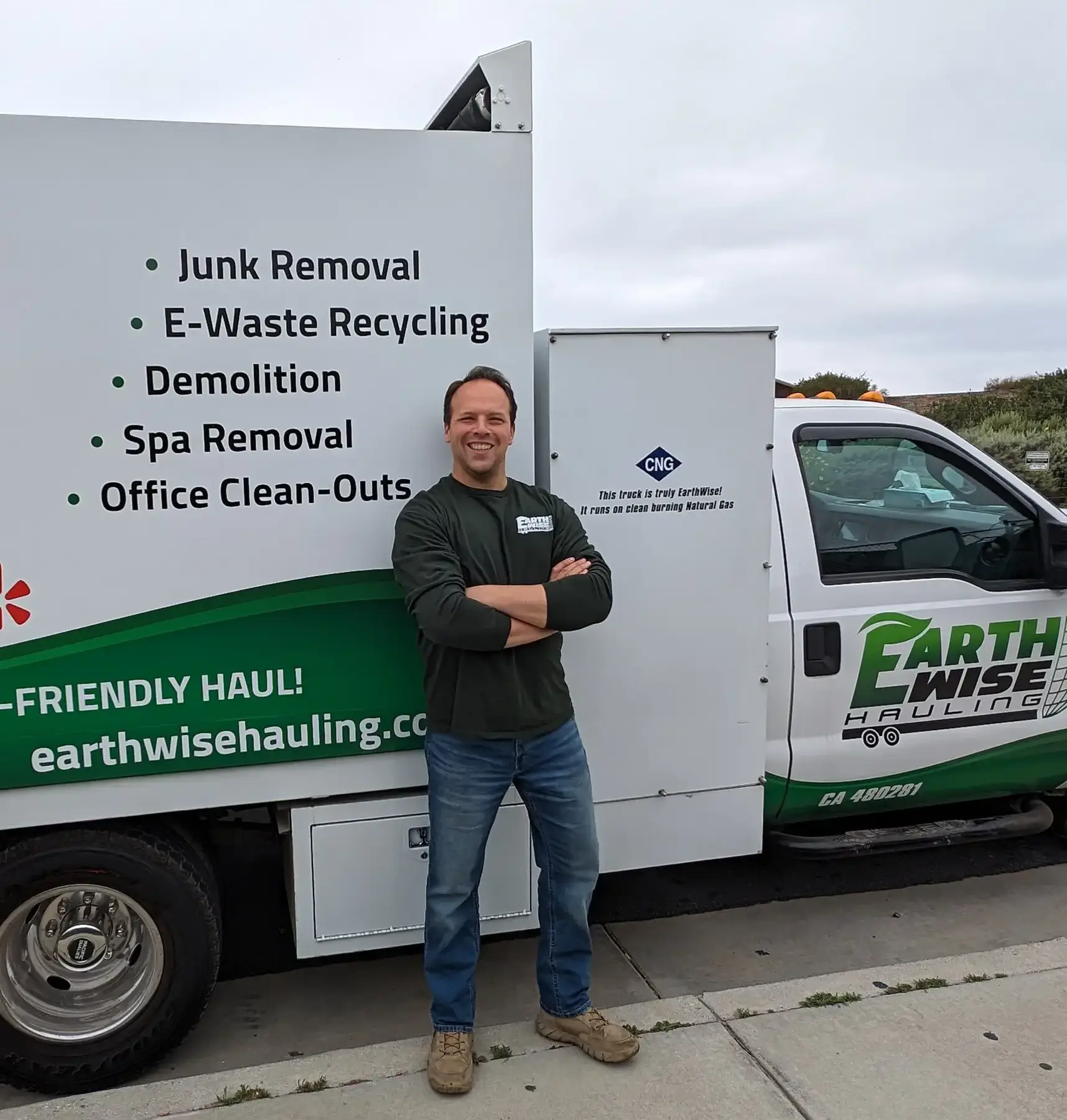 Owner, Joe in a EarthWise shirt stands with crossed arms in front of a white hauling truck showing services: Junk Removal, E-Waste Recycling, Demolition, Spa Removal, Office Clean-Outs (Earth Wise Hauling).